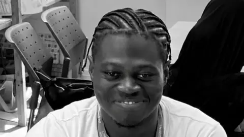 Umar Dumbuya is looking at the camera with a smile. He is wearing a white t-shirt and braided hair