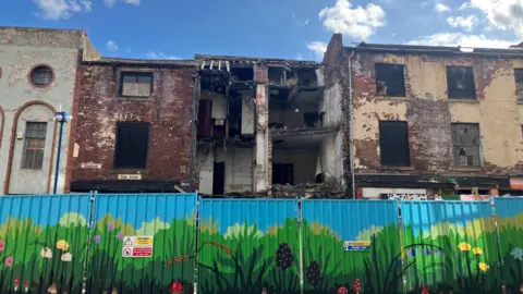 A derelict building is fenced off. The interior of the building is visible in the photo as the frontage has fallen away.