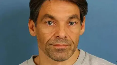 Robert Brown looks directly at the camera for a police mugshot. He has short, dark brown hair and a short shaved beard. He also has blue eyes and is wearing a pale grey sweatshirt