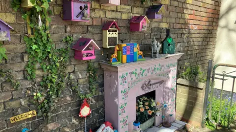 Shariqua Ahmed/BBC A pink fire place with colourful books on it. Several small wooden bird houses have been stuck on a stone wall behind it.