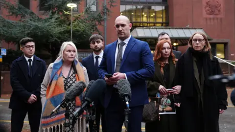PA Media Detective Inspector Joe Davenport stands with members of Leo Ross' foster family outside Birmingham Crown Court. There are three women dressed in winter coats stood with four men in dark clothing outside the court. Their faces are sullen as Davenport speaks to the media. The court can be seen behind the group.