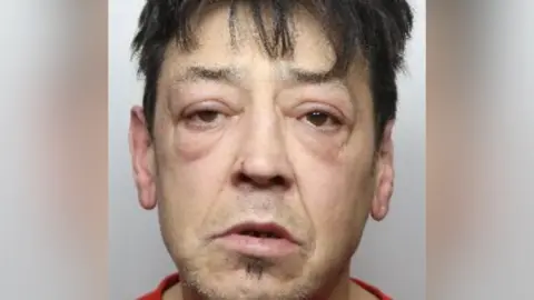 A police mugshot of a dishevelled looking man. He has eye bags and puffy eyelids. He has a bit of hair below his mouth but is mostly clean shaven. He has messy black hair around his forehead.