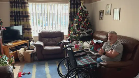 Neville Bradford sits on his living room sofa to right of camera. He is talking to someone out of frame to the left. Wearing a grey polo top and blue bottoms. It is clear he is a double leg amputee. A black wheelchair with a tartan blanket on the seat is in front of him, There is a Christmas tree in one corner behind him next to the window.