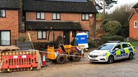 BBC A brick house with a marked yellow and blue fluorescent police car parked outside. There is also construction equipment outside the front of the property with cones and tape. 