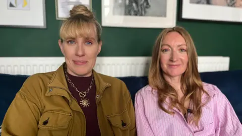 Nicola and Lowri sat side by side on a dark blue sofa. Nicola has a blonde fringe and wears a khaki jacket with a purple top and a large sun necklace pendant. Lowri wears a pink shirt. 