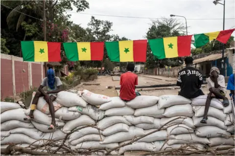 MUHAMADOU BITTAYE/ AFP Protesters sitting on sandbag barricades under Senegalese flags.