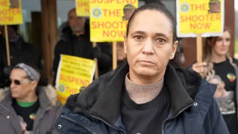 Selina Freckleton stands at the protest. There are people with yellow placards behind her. She is wearing a beige polo neck and black T-shirt with her nephew's picture on it and a navy coat.