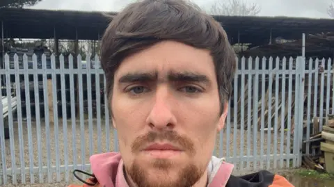 Luke Jones has brown hair and eyebrows and is standing in front of an iron gate.