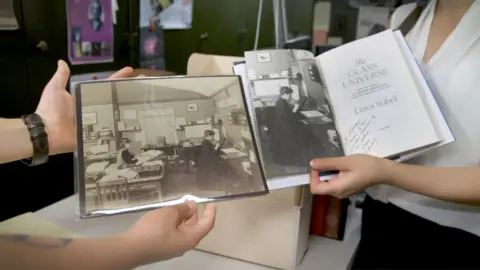 Alex Newman/PRI Bouquin, left, and Smith Zrull, right, hold up an original image of Williamina Fleming posing in the plate stacks in a 1891 photo that was the first photo used in bestselling author Dava Sobell's 2016 book, "The Glass Universe." Smith Zrull says they know the 1891 image is posed because a window is closed and the tool Fleming is using to study a plate only works with window light.