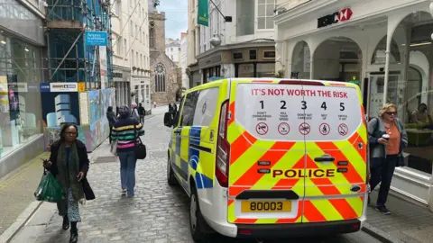 John Fernandez/BBC Passers-by are walking past a pole van parked up in a narrow street in Guernsey with shops on either side and an HSBC bank on the right. One woman is holding a drinks cup. There is a church tower at the end of the street.