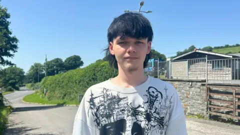 Conor, wearing a white t shirt with a black cross on it. He has a black mullet and short stubble. There is a winding road and a building behind him.