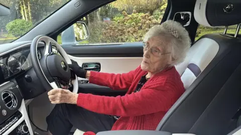 A woman with white hair, glasses and a red cardigan sitting in a car, holding the steering wheel in both hands
