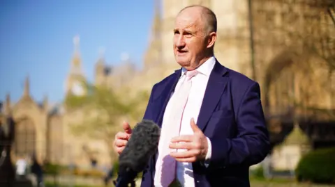 James Manning/PA Wire Kevin Hollinrake, the Conservative Party chairman, is standing outside Westminster, talking to a reporter. He is wearing a purple suit jacket with a pink shirt and tie.