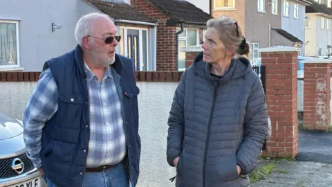 Kevan Hill, wearing a blue checked shirt and blue gilet, chatting to Ronda, right, wearing a blue coat. They are outside standing on the street, with houses in the background