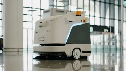 Getty Images A white sweeping robot cleans the tiled floors of a large hall. The robot is square shaped and has a dark coloured side with a blue light. It is on wheels and has black brushes which clean the floor.