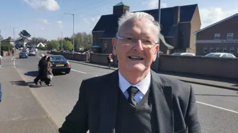 Sydney Murphy is standing on a street. He wears glasses. He has a black suit on, a black tie and jumper and white shirt. People are crossing the road behind him. Buildings belonging to St Paul's Parish are behind him.