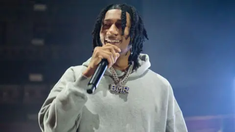 Digga D on stage wearing a grey hoodie and a necklace holding a black microphone to his mouth. He has long, braided black hair.