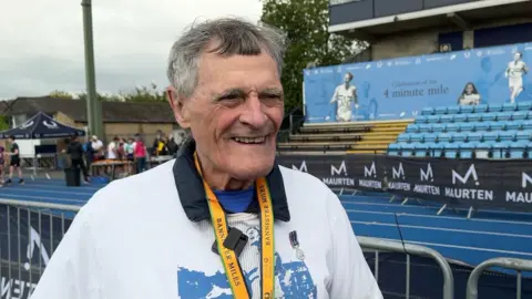 A man wearing a white jumper and Banister Miles medal smiles in front of a blue running track.
