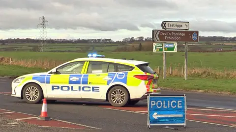 A police car sits behind a diversion sign with a road sign for Eastriggs in the background and fields with an electricity pylon in the distance