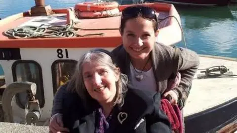 Pippa Marshall Woman in a wheelchair with grey hair smiling with her daughter holding on to the wheelchair behind her with fishing boat and harbour behind