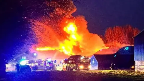 Orange flame and smoke erupts from a farm building. The outline of fire engines can be seen in the foreground.