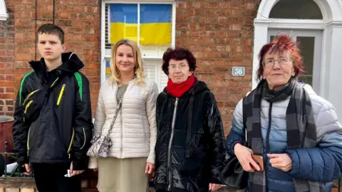 Taras, Nataliya, Helen and Iren standing outside a house near where they live with a Ukraine flag in the window