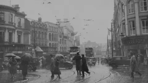 Fox Photos/Getty Images/Hulton Archive A black and white archive photo of a wet and windy day in Castle Place in Belfast city centre, circa 1925.  There are tall, ornate buildings lining the cobbled street.  Several pedestrians are walking in the rain, some are holding umbrellas.  Motor cars and double decker trams are travelling along the street.  There is a large union flag flying from one of the buildings.