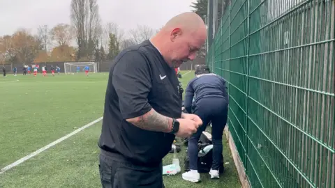 Warren Muggleton/BBC A side-on profile of Stuart Nixon in a black referee kit, standing beside an all-weather green football pitch. He appears to be fiddling with something with both hands, looking downward.