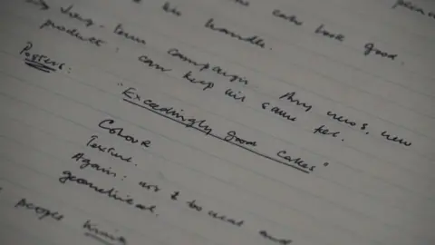 Shaun Whitmore/BBC A close-up of a page from a lined notebook with handwritten notes. It references the tagline exceedingly good cakes. 