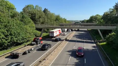 Steve Hubbard/BBC Dual carriageway showing traffic including lorries, vans and cars heading up from the bottom left of the photo. The vehicles are going under two motorway bridges. A police van is parked on the most distant bridge. A red van is visible heading in the opposite direction. There are trees on either side of the road.
