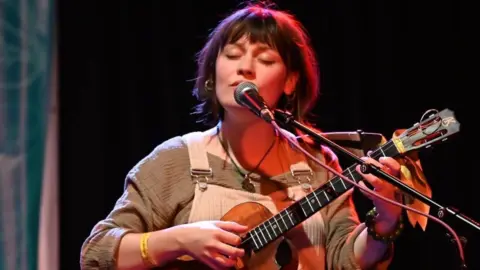 Peter Brown A woman with dark hair with her eyes closed sings into a microphone while playing a ukulele