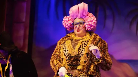 Dan Baugh Pantomime dame Joe Meloy dressed in a yellow dress and a pink wig, wearing glasses and lipstick. 