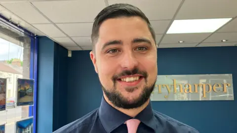 Sean with short black hair and a short beard, wearing a navy blue shirt and pink tie, standing in an estate agent's office