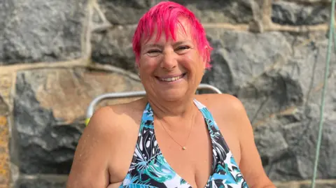 Janine Le Cras has bright pink hair and is sitting on a camping chair. She's wearing a blue patterned swimsuit.