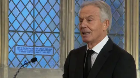 Former PM Tony Blair with gray hair mid-spit because he delivers a ecstasy for John Prescott. There is a microphone in front of him. He is wearing a black suit and tie