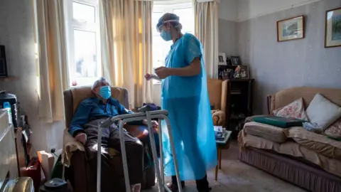 A healthcare worker standing and dressed in a blue gown chats to a housebound patient in his front room. The patient is sat down on a chair with a medical walking frame in front of him. He is wearing a mask. There is a sofa on the right of the picture and sun is coming through the windows behind.