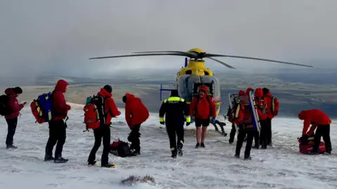 A team of Keswick Mountain Rescue volunteers, who are all wearing red waterproofs and large rucksacks, stand on top of the summit of Blencathra. There is a yellow helicopter behind them.