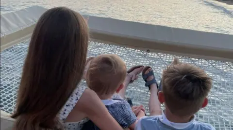 Rebecca Saunders Mrs Saunders' three children sit with their backs to the camera overlooking the sunset over the see. They are sat on a large white net, directly over the water. 