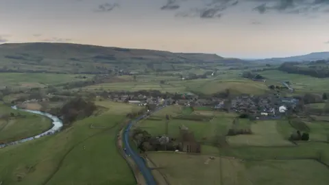 This is an overhead shot of Bainbridge, in the Yorkshire Dales. A river is winding through the left of the shot, the village is on the right, the hills are in the background, a dark and threatening sky above. North Yorkshire Council says the size - and rural nature - of the area it serves adds extra costs to its services.