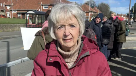 Joan Dennis stands at the side of the road with a concerned expression on her face. She is wearing a salmon-coloured jumpers and a pink coat. She has short grey cropped hair. 