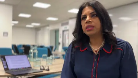BBC A woman in a navy dress with red piping around the collar and buttons speaks to someone off camera. She is in a room with desks and blue chairs. An open laptop can be seen on a table to her left.