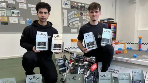 Two students, Adil and Kacper, from St Mary Redcliffe and Temple sixth form centre are posing for the photo. They are on one knee with trophies from the various VEX Robot Championships they have competed in. Adil has black curly hair and Kacper has brown hair, they are both wearing black clothes. 