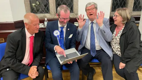 L-R: Dave Curtis, Malcolm Ferey, Kevin Lewis and Suzie Webb. All are looking at a laptop which Ferey is holding while they sit down on blue cushioned chairs. Lewis is holding both his hands up and has his mouth wide open.