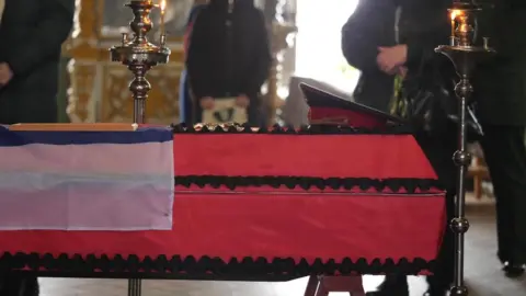 BBC A coffin in red material covering with a serviceman's cap laid on top