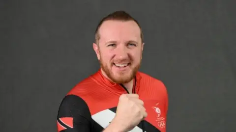 Getty Images Axel Brown, a young man wearing a red, black and white Olympic snowsuit. He has short dark hair and a beard.