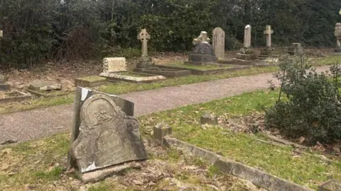 BBC/ Alex James In the foreground of the image there is a grave that has been split in half, with the top half resting against the bottom. Between the foreground and background there is a path. In the background there is a row of graves with two also broken. 
