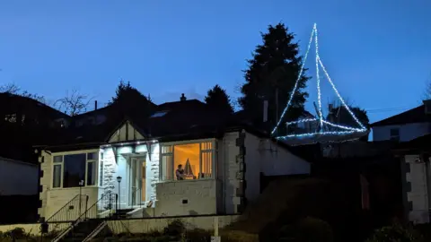 A photo of Ian's house with the illuminated yacht rising up behind it shining against the night sky. The house is a white brick detached house. The front room light is on and you can see a figure holding a small model yacht