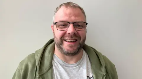 A man in glasses, with a short beard and moustache smiles at the camera. He is wearing a green hoodie over a grey T-shirt
