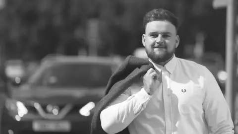 Family Handout A black and white photo of Luke, smiling to camera, looking smart in a white shirt and holding a dark jacket over his shoulder