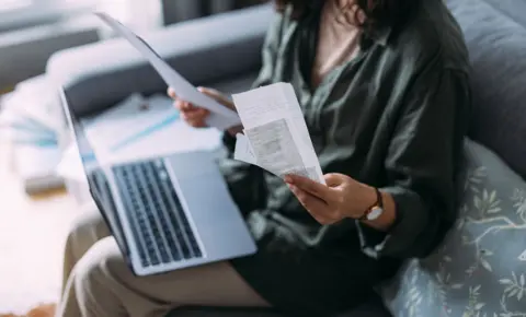 Getty Images Woman overwhelmed with bills to pay, she is sitting on her sofa with a laptop holding receipts